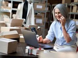 Senior, female business owner works on laptop and takes a phone call in shipping warehouse